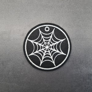 Puede incluir: Un parche bordado en blanco y negro con un diseño de telaraña. El parche es circular y tiene un contorno blanco.
