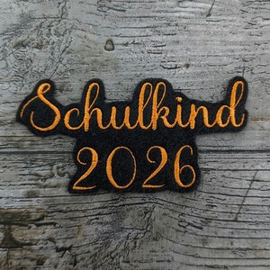 以下が含まれることがあります： オレンジ色の筆記体で「Schulkind 2026」と書かれた黒色の刺繍パッチ。 パッチは角が丸い長方形で、風化した灰色の木製の背景に配置されています。