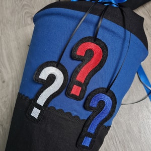 Puede incluir: Un cono de tela azul y negro con tres parches de tela en forma de signo de interrogación en rojo, blanco y azul.