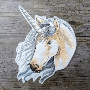 Peut inclure: Patch brodé représentant la tête d'une licorne blanche avec une corne dorée et une crinière bleu et blanche. La licorne est tournée vers la gauche et a un œil fermé.