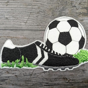 Op de afbeelding: Een zwart-witte voetbal met een zwart-witte voetbalschoen met groen gras op een grijze houten achtergrond.