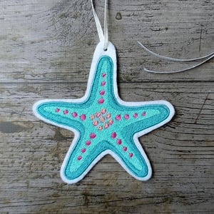 May include: A teal blue starfish ornament with pink and yellow accents. The ornament is made of felt and has a white ribbon attached to the top.