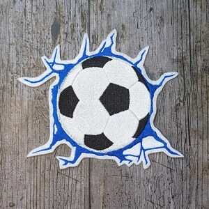 Könnte beinhalten: Ein weißer und schwarzer Fußball mit blauen Nähten ist in einem weißen und blauen gestickten Patch zu sehen. Der Patch ist so gestaltet, dass es aussieht, als würde der Ball eine Oberfläche durchbrechen.