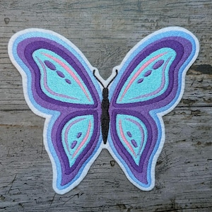 Peut inclure: Écusson papillon brodé dans les tons de violet, bleu et rose. Le papillon a un design en couches avec une bordure blanche. Les ailes présentent un centre bleu clair avec des accents roses et un contour violet foncé.