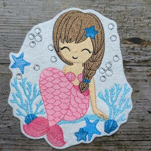 May include: Embroidered patch of a smiling mermaid with brown hair and a pink tail. The mermaid is surrounded by blue starfish, coral, and bubbles on a white background. A cute design for apparel or accessories.