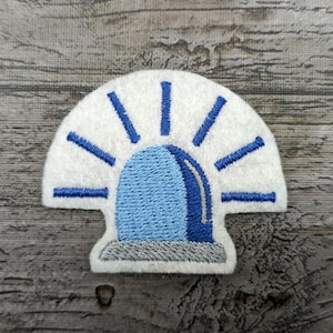 May include: A white felt patch with a blue and gray embroidered police siren. The siren has a white light around it and blue and white rays coming out from the top.