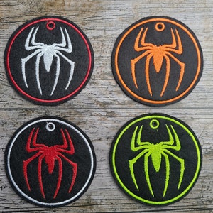 Peut inclure: Quatre patchs ronds en feutre noir avec des araignées blanches, orange, rouges et vertes brodées. Chaque patch a un petit anneau argenté en haut.