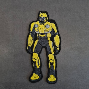 Puede incluir: Parche bordado amarillo y negro de un personaje robot. El robot tiene un casco amarillo con una visera negra y un cuerpo negro con detalles amarillos.