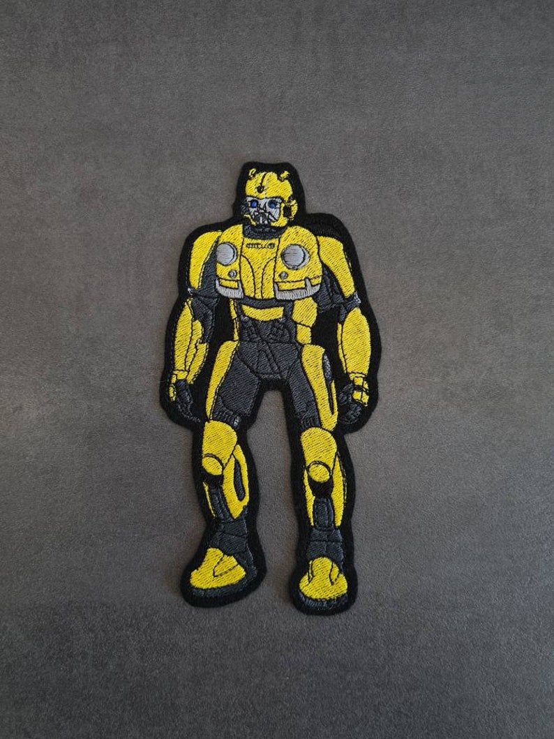 May include: Yellow and black embroidered patch of a robot character. The robot has a yellow helmet with a black visor and a black body with yellow accents.