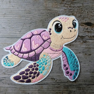 May include: Embroidered patch of a cartoon sea turtle. The turtle features a light tan face with large black eyes and a smiling mouth. The shell is purple and pink, and the flippers are blue, purple, and pink. The patch is on a wooden surface.