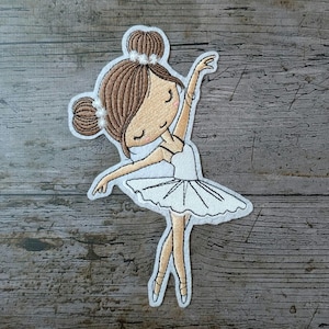 Puede incluir: Parche bordado con una bailarina de dibujos animados en una pose elegante. La bailarina tiene el pelo castaño peinado en dos moños y lleva un tutú blanco y zapatillas de punta. El parche está contorneado en blanco, sobre un fondo de madera.