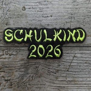 Könnte beinhalten: Ein schwarzer, gestickter Aufnäher mit den Worten "SCHULKIND 2026" in neongrüner Schrift. Der Aufnäher ist rechteckig mit leicht unebenen Kanten, und der Text ist in einer stilisierten Schriftart gehalten. Der Hintergrund ist eine verwitterte Holzoberfläche.