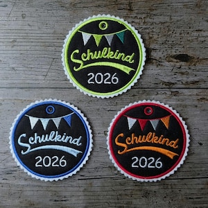 Könnte beinhalten: Drei gestickte Aufnäher mit dem Text "Schulkind 2026". Jeder Aufnäher ist kreisförmig mit schwarzem Hintergrund, einem gewellten Rand und einem Bannerdesign. Die Aufnäher haben grüne, blaue und rote Ränder.