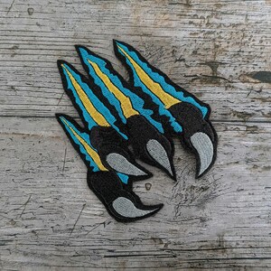 Könnte beinhalten: Gestickte Krallen-Patches mit schwarzer Basis, blauen und gelben Akzenten und grauen Spitzen. Die Patches sind auf einer verwitterten Holzoberfläche angeordnet. Ideal zum Individualisieren von Kleidung oder Accessoires, diese Patches verleihen eine einzigartige Note.