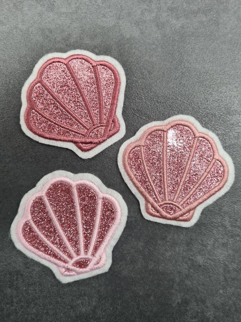 May include: Three pink glitter seashells with white outlines. The seashells are embroidered and have a sparkly finish.