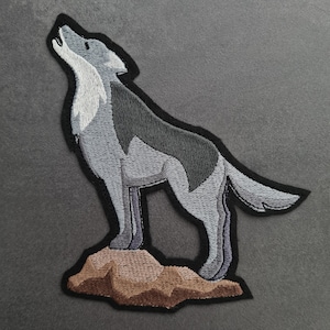 Może przedstawiać: Naszywka haftowana do prasowania przedstawiająca szarego wilka wyjącego do księżyca, stojącego na brązowej skale.