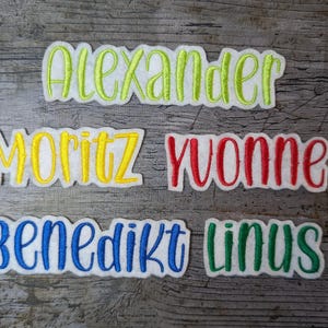 Könnte beinhalten: Fünf bestickte Namensschilder auf einem Holzgrund. Die Namen sind Alexander, Moritz, Yvonne, Benedikt und Linus. Die Schilder sind in verschiedenen Farben, darunter Gelb, Grün, Rot, Blau und Weiß.