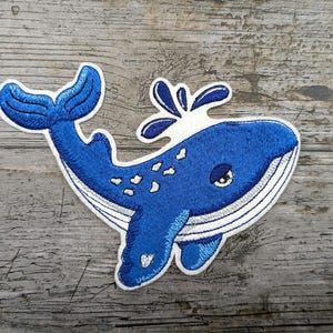 Può includere: Toppa ricamata a forma di balena blu con cuciture bianche, pancia bianca e dettaglio di spruzzi d'acqua. La balena ha un occhio chiuso e una piccola bocca.
