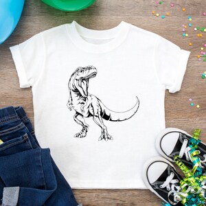 May include: A white t-shirt with a black and white illustration of a dinosaur on the front. The shirt is laying on a wooden surface with blue jeans, a blue balloon, a green balloon, and confetti.