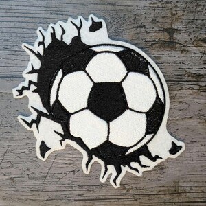 Puede incluir: Un parche bordado en blanco y negro de un balón de fútbol rompiendo una pared. El balón de fútbol tiene un diseño clásico con costuras negras.