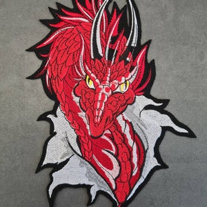 May include: An embroidered patch featuring a detailed red dragon head with sharp white horns and piercing yellow eyes. The dragon is depicted emerging from a torn white fabric effect, outlined in black thread against a neutral grey background.