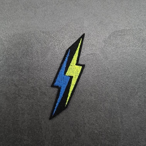 Puede incluir: Un parche bordado con forma de rayo amarillo y azul. El parche es negro con un contorno amarillo y un rayo azul en su interior.