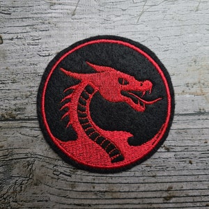 Może przedstawiać: Czarny okrągły znaczek z haftowanym wzorem czerwonej głowy smoka.