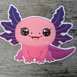 Pode incluir: Um patch de axolotl rosa e roxo com um rosto sorridente e olhos grandes e expressivos. O axolotl tem brânquias roxas e uma cauda a condizer. O patch é feito de tecido e tem uma borda branca.
