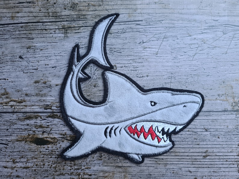 May include: A white felt shark patch with black outline and red teeth. The shark is facing right and has a large open mouth.