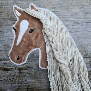 Op de afbeelding: Een bruine paardenkop-applicatie met een witte ster op het voorhoofd en een lange, gevlochten, crèmekleurige manen.