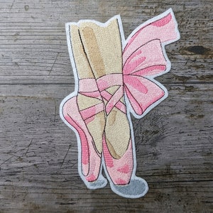 May include: Embroidered patch featuring a pair of pink ballet pointe shoes with ribbons tied in a bow. The shoes are a light tan color with pink accents and are set against a wooden background.