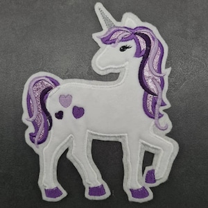 Może przedstawiać: Haftowana naszywka z białym jednorożcem z fioletową grzywą, ogonem i kopytami. Jednorożec ma srebrny róg i trzy fioletowe plamki w kształcie serca z boku. Naszywka jest obramowana na biało.