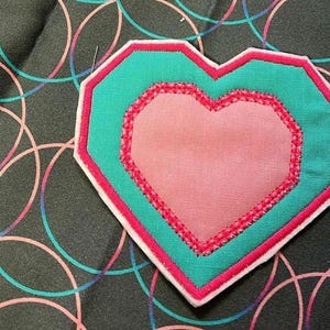May include: A pink heart-shaped patch with a turquoise border and white stitching. The patch is on a black background with a pattern of overlapping circles.