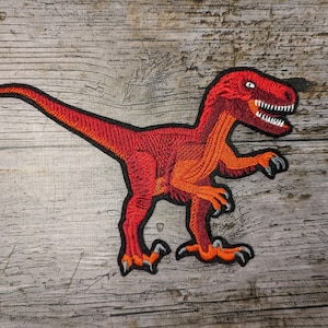 May include: Embroidered patch featuring a red and orange dinosaur, likely a velociraptor, with sharp teeth and claws. The patch has a black border and is set against a weathered, gray wooden background. A fun accessory for clothing or bags.