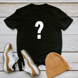 May include: A black t-shirt with a white question mark on the front, a pair of white and gold sneakers, and a brown beanie.