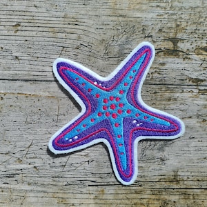 Puede incluir: Una estrella de mar azul y morada con lunares rosas, bordada en un parche de tela blanca.