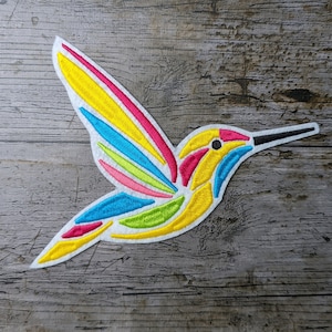 Puede incluir: Un parche bordado de colibrí colorido con acentos amarillos, rosados, azules y verdes. El parche está sobre un fondo de madera.