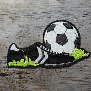 Op de afbeelding: Geborduurde patch met een voetbal en een voetbalschoen. De bal is zwart en wit, en de schoen is zwart met witte strepen. Groen gras bevindt zich aan de basis van de schoen en de bal. De patch bevindt zich op een houten achtergrond.