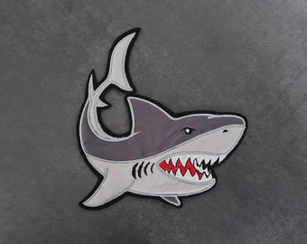 Puede incluir: Un parche de tiburón bordado en gris y blanco con una boca abierta roja y dientes afilados. El tiburón está mirando hacia la derecha y tiene una aleta blanca.