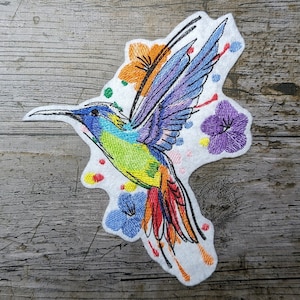 May include: Embroidered patch featuring a colorful hummingbird with a long, red tail, surrounded by flowers in shades of purple, blue, orange, and green. The patch is on a white background.