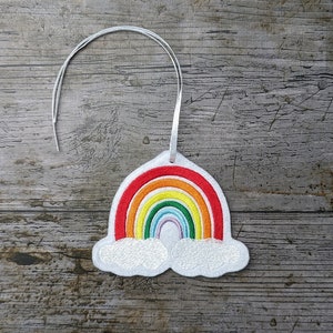 Può includere: Un ornamento arcobaleno in feltro bianco con uno spago argentato per appenderlo. L'arcobaleno ha strisce rosse, arancioni, gialle, verdi, blu, indaco e viola. L'arcobaleno è circondato da due nuvole bianche.