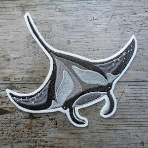 May include: Embroidered patch featuring a manta ray design. The patch has a white border and showcases shades of gray and black, with subtle details. The manta ray's wings are spread, and the design is on a wooden surface.