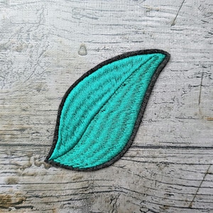 Può includere: Una toppa ricamata a forma di foglia con un vivace centro color verde acqua caratterizzato da linee di cucitura dettagliate, circondata da un bordo nero solido. La toppa presenta un design curvo e appuntito che imita la forma naturale di una foglia.