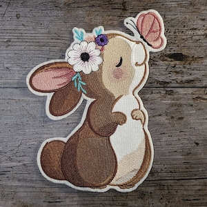 Pode incluir: Etiqueta bordada de um coelho marrom com nariz rosa e pêlo branco, usando uma coroa de flores azuis e roxas. O coelho está olhando para cima para uma borboleta rosa.