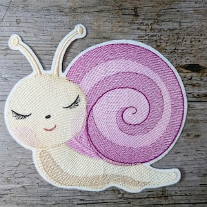 Puede incluir: Un caracol bordado de color rosa y blanco con un diseño de ojo cerrado. El caracol está sobre un fondo de madera.