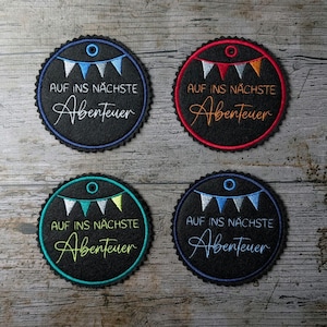 Op de afbeelding: Vier ronde patches met geschulpte randen. Elke patch is zwart met een gekleurde rand en heeft de tekst "AUF INS NÄCHSTE Abenteuer" in witte letters. Boven de tekst staat een banner met kleine vlaggetjes.