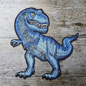 Könnte beinhalten: Ein blauer gestickter Aufnäher mit einem Tyrannosaurus Rex Dinosaurier. Der Dinosaurier steht auf seinen Hinterbeinen, mit offenem Maul und sichtbaren Zähnen. Der Aufnäher befindet sich auf einem hölzernen Untergrund.