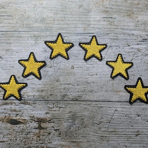 Puede incluir: Seis parches de estrellas amarillas con bordes cosidos en negro dispuestos en arco. Los parches están hechos de tela texturizada y están colocados sobre un fondo de madera envejecida. Cada estrella tiene cinco puntas.