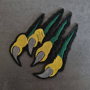 May include: A black, green, and yellow embroidered patch of a claw with sharp points.