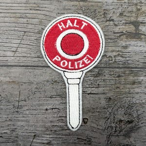 Puede incluir: Parche bordado blanco y rojo con las palabras "HALT POLIZEI" y un círculo rojo en el centro. El parche está unido a un palo blanco.